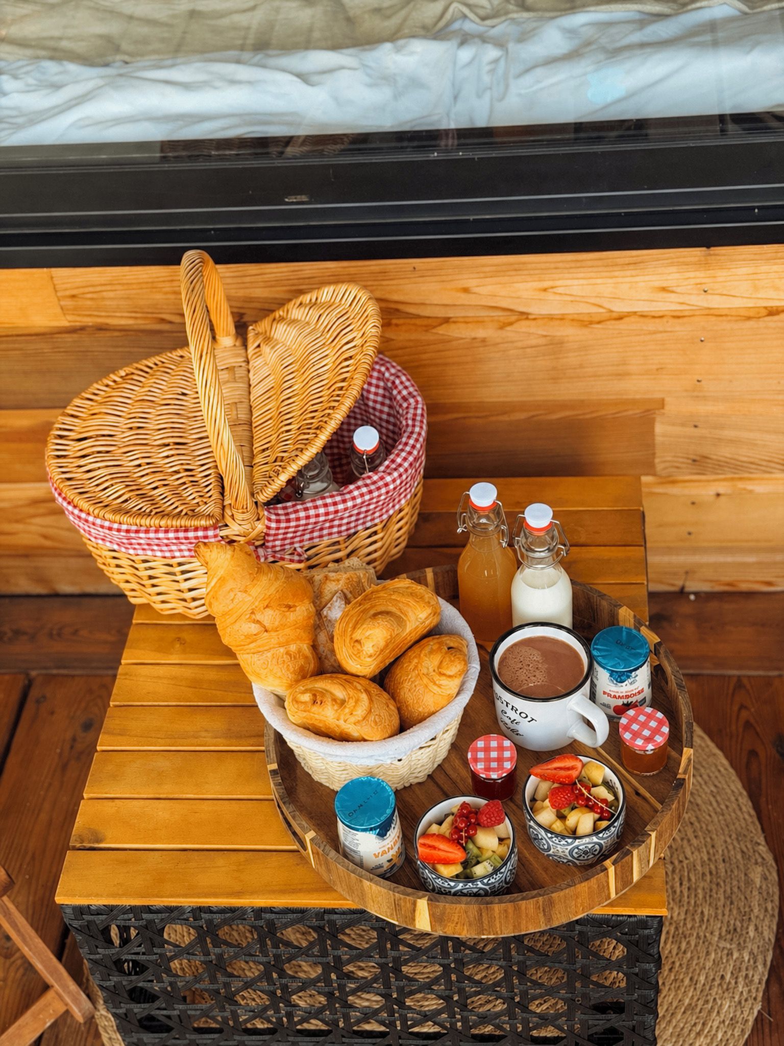 A wooden table with a wicker basket containing croissants, a bottle of milk, a mug of coffee, and bowls of fruit.