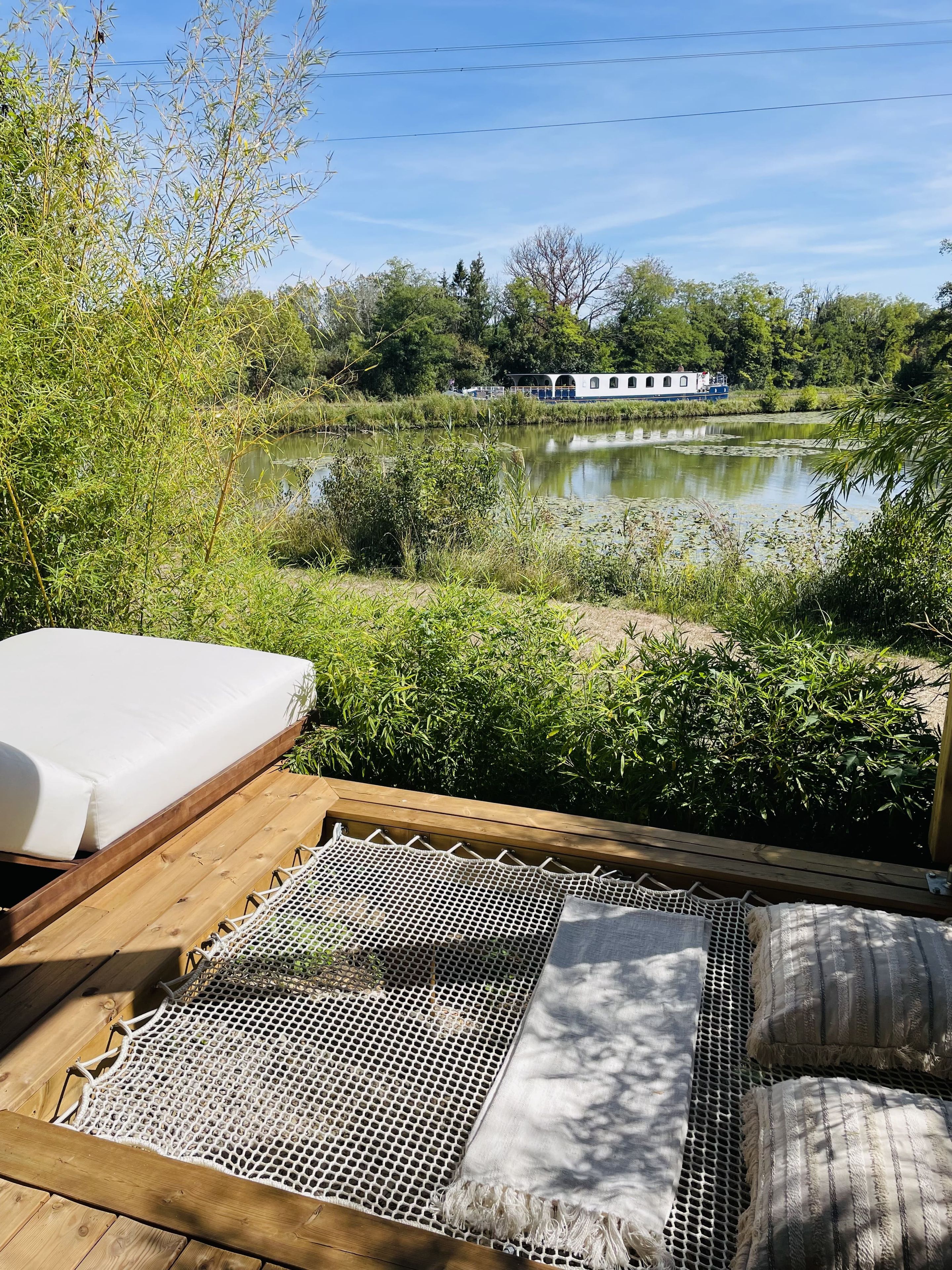 En træterrasse med hængekøje og madras, med udsigt over en rolig sø og en husbåd.