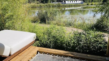 En træterrasse med hængekøje og madras, med udsigt over en rolig sø og en husbåd.