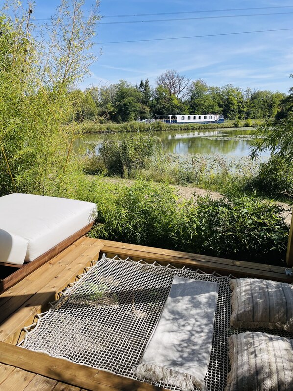 A wooden deck with a hammock and a mattress, overlooking a calm pond and a houseboat.