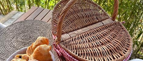 A wicker basket with a handle on a wooden table, a bowl of pastries, and a woven placemat.