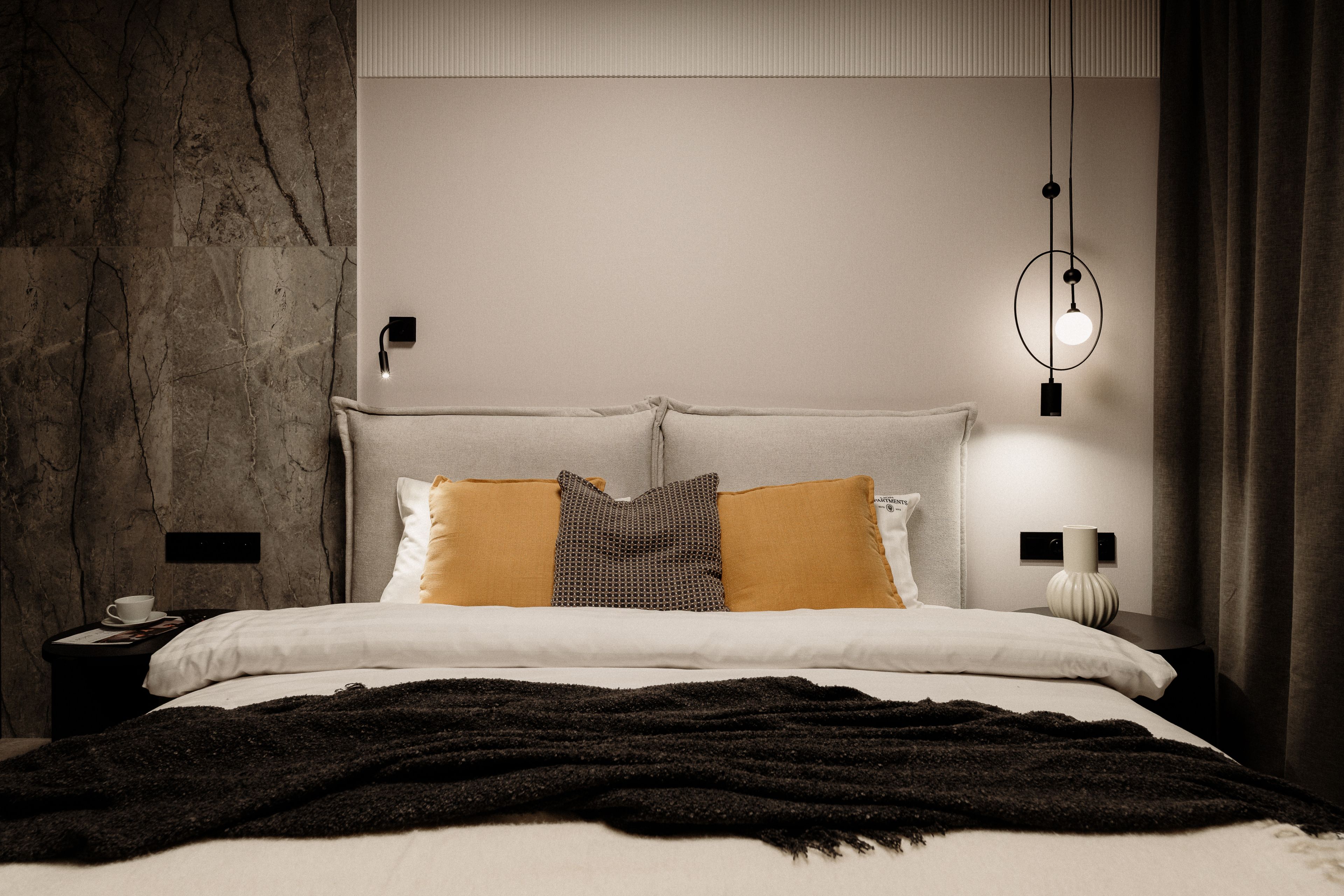 A modern bedroom with a stone wall, a bed with multiple pillows, a bedside table with a lamp, and a hanging light fixture.