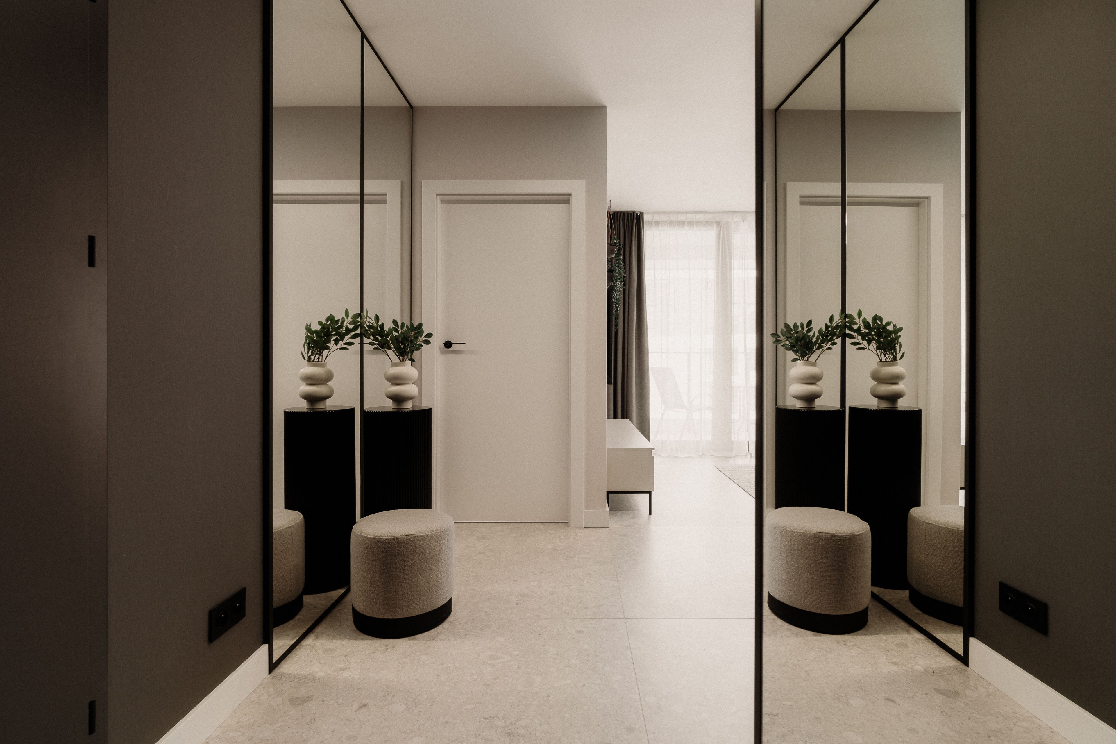A hallway with a mirrored wall, a door, a bench, and two round ottomans.
