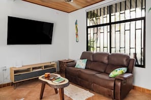 Ein Wohnzimmer mit braunem Ledersofa, einem flachbildschirmfernseher an der Wand, einem Holztisch und einem Fenster mit Sprossen.