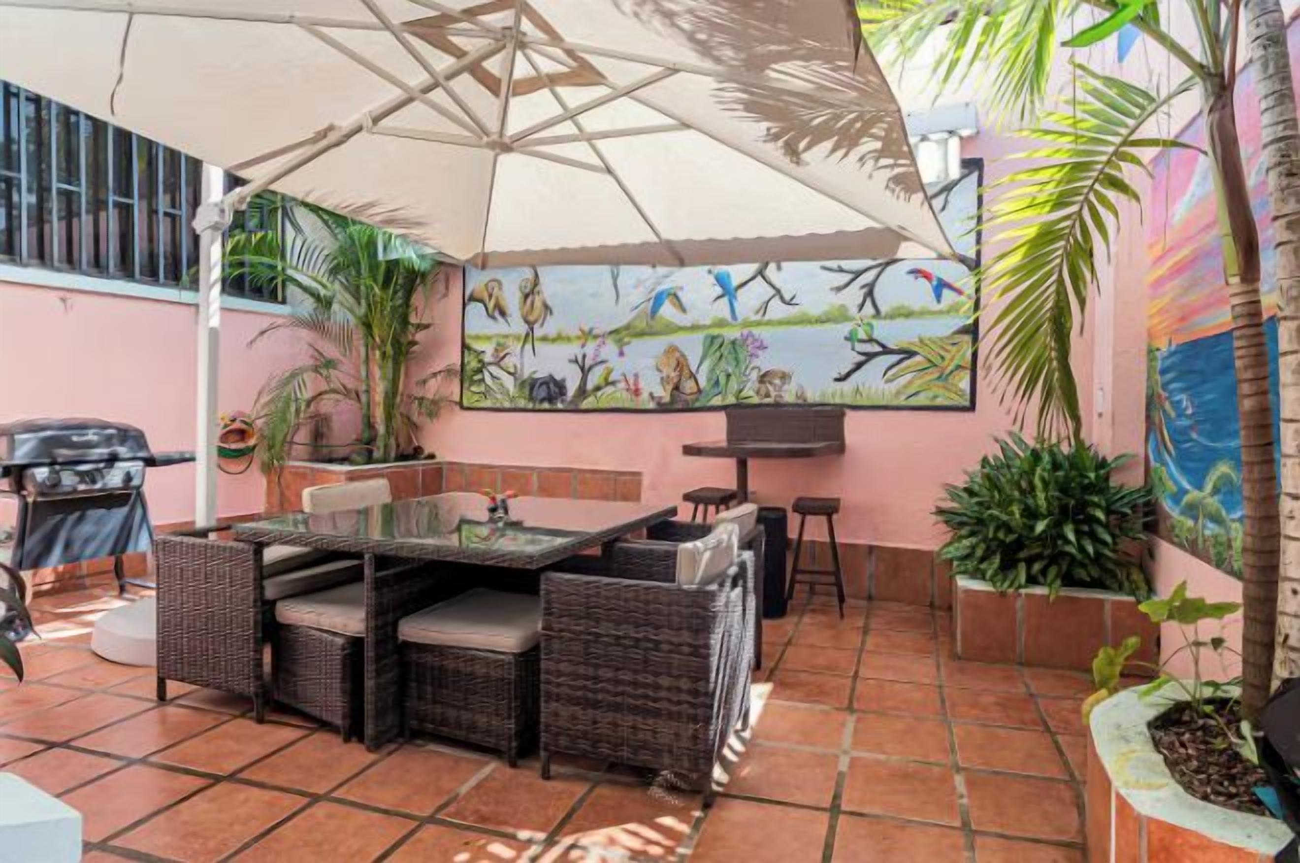 An outdoor patio with a dining table, chairs, and a large umbrella. There are potted plants and a mural on the wall.