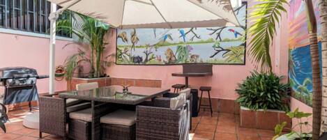 An outdoor patio with a dining table, chairs, and a large umbrella. There are potted plants and a mural on the wall.