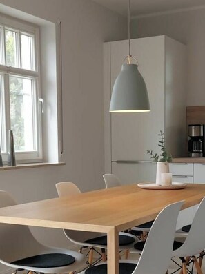 Eine moderne Küche mit einem Holztisch, weißen Stühlen, einer grauen Pendelleuchte und einer Kaffeemaschine.
