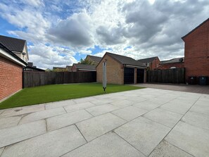 A residential backyard with a paved patio, a grassy lawn, and a brick house.