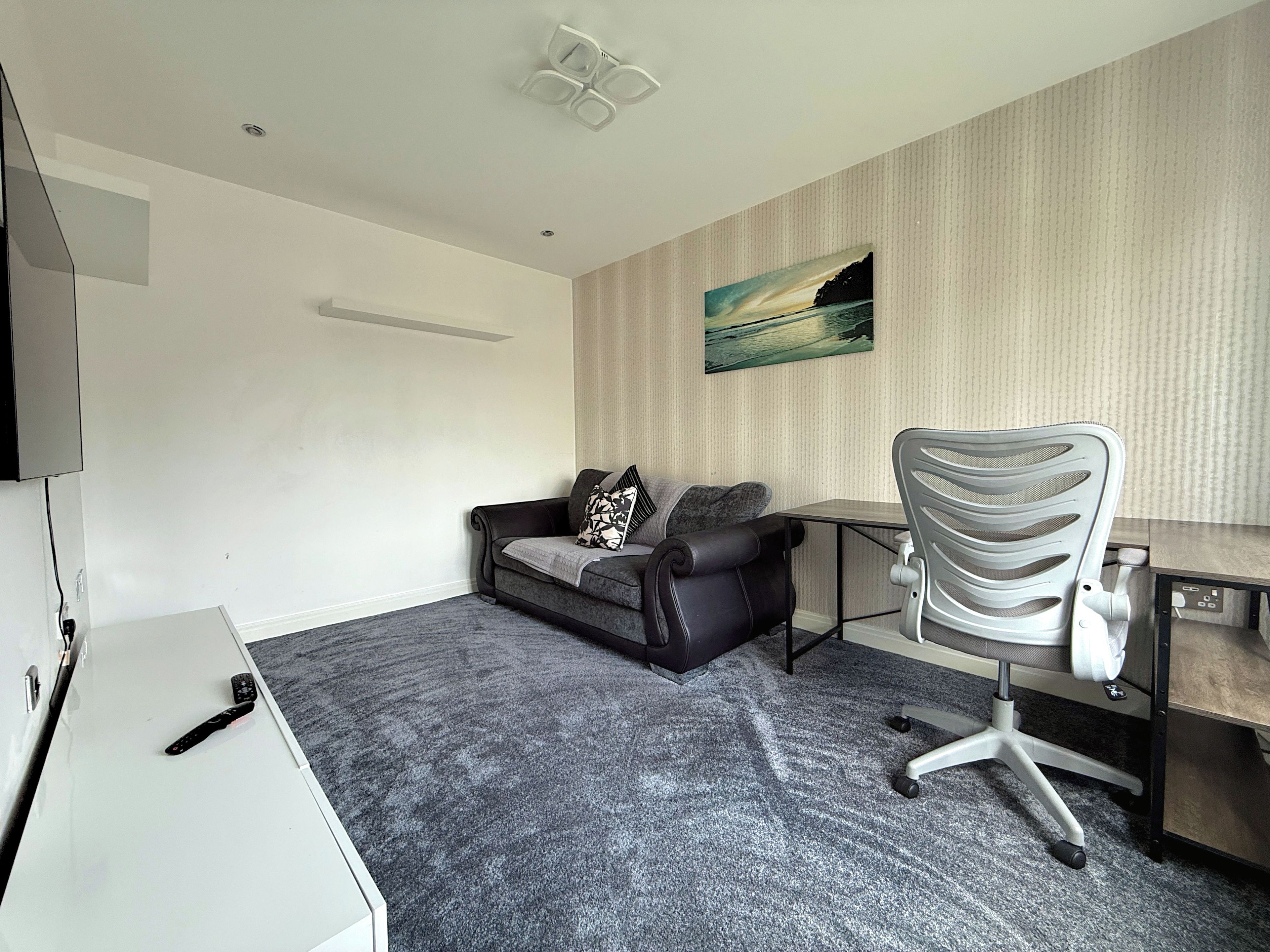 A room with a grey carpet, a black leather sofa with a patterned pillow, a white desk, and a grey office chair.