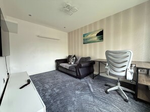 A room with a grey carpet, a black leather sofa with a patterned pillow, a white desk, and a grey office chair.