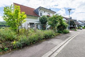 A residential street with a white house featuring a red-tiled roof, a garden with various plants, and a clear sky.