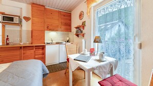 Een knus ingerichte kamer met een bed, een tafel met lamp, een keukenhoek en een raam met kantgordijnen.