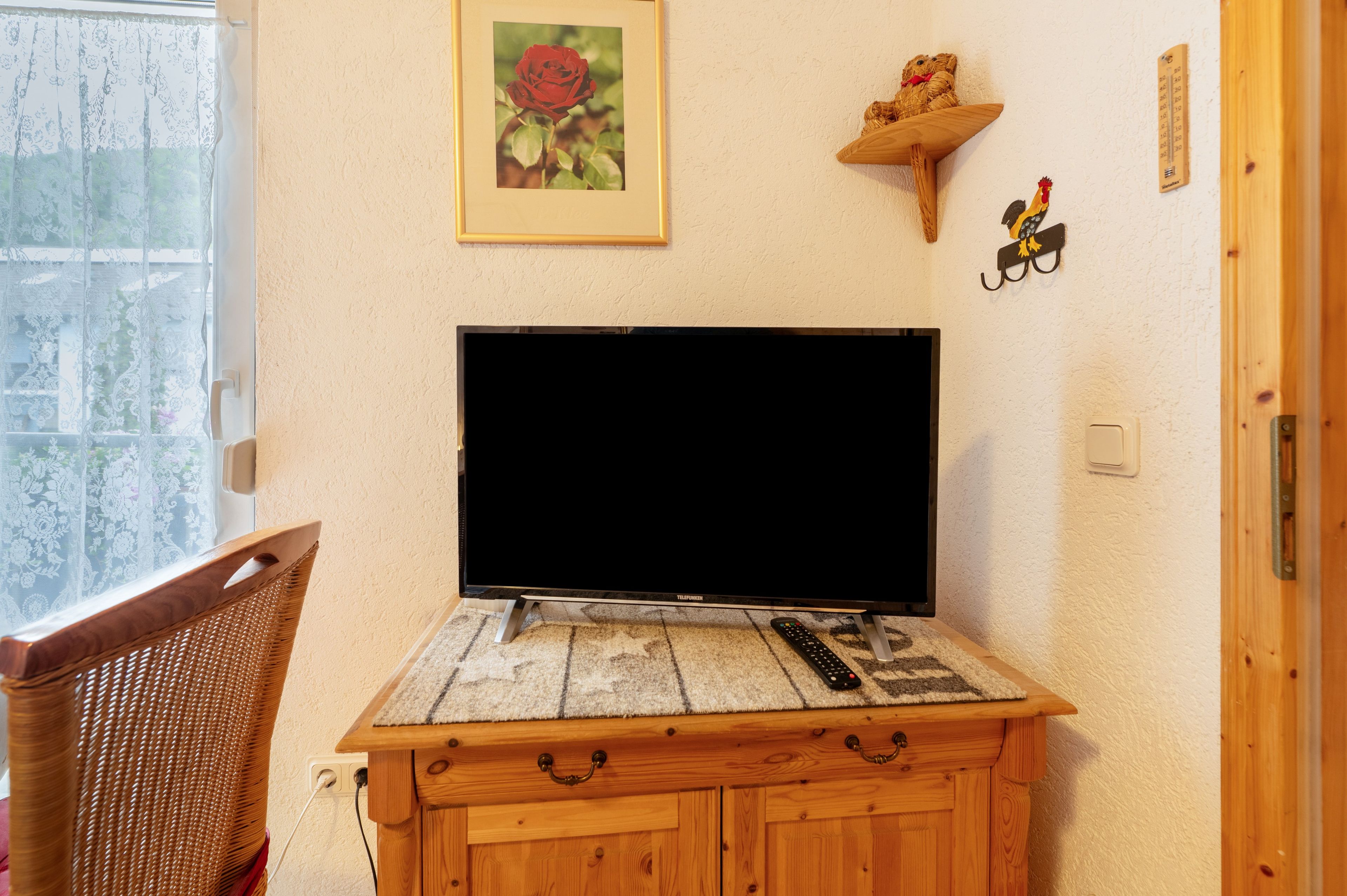 Ein Fernseher auf einem Holzständer mit Fernbedienung, ein Korbstuhl, ein gerahmtes Rosenbild und ein an der Wand befestigtes Thermometer.