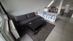 A compact room with a sofa, a small table, a bed with a checkered bedspread, a kitchenette, and a window with blinds.