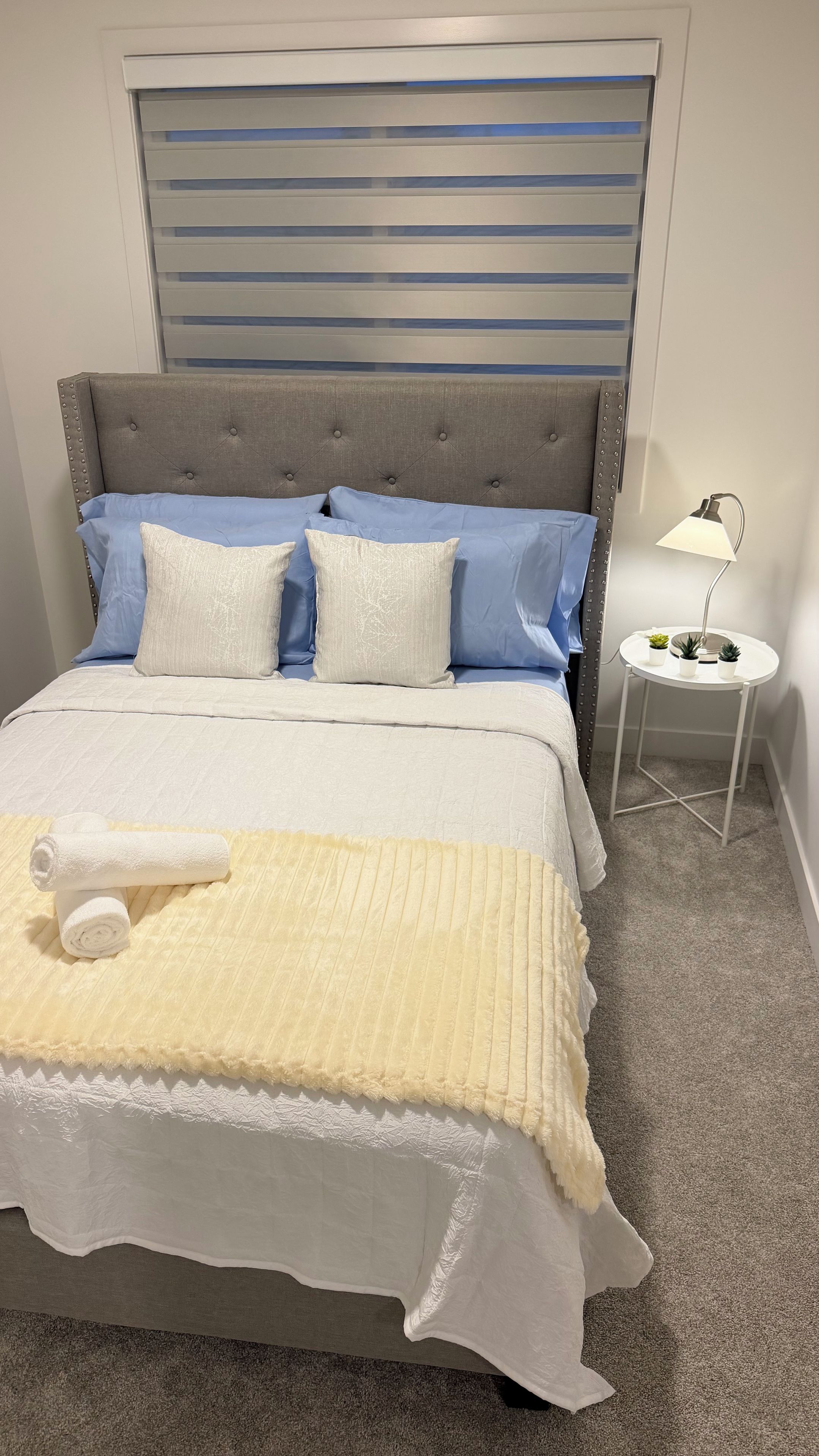 Una cama bien hecha con cabecera gris, una manta blanca y amarilla, y una toalla enrollada. Hay una mesita de noche pequeña y blanca con una lámpara y algunas plantas en macetas.