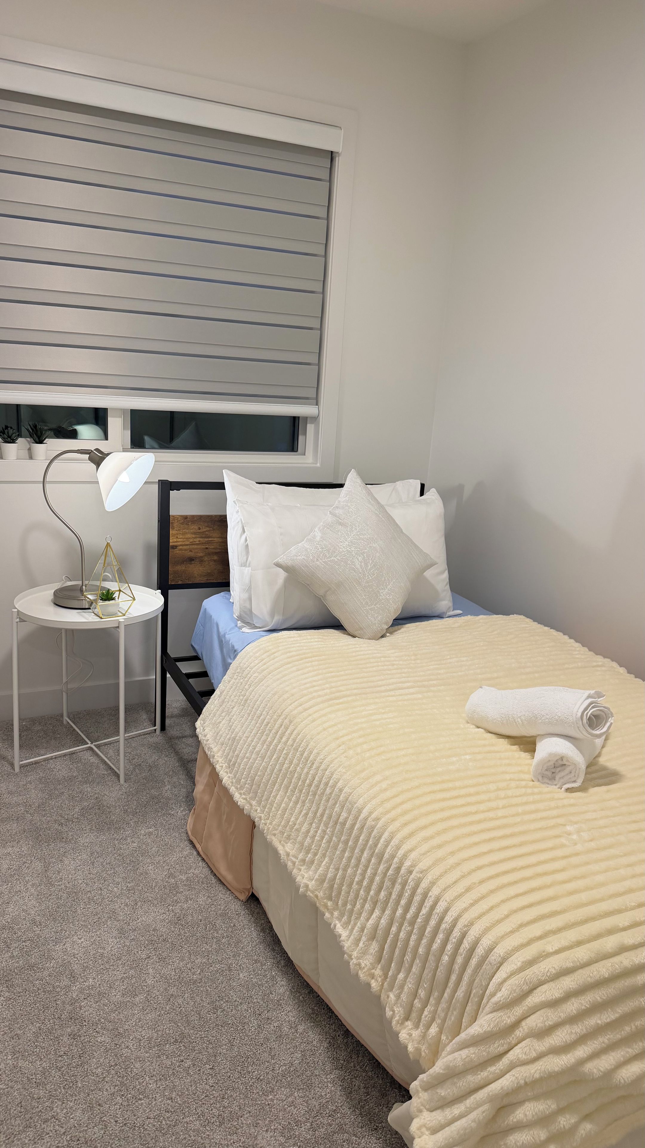 A bedroom with a bed, a bedside table with a lamp, a window with blinds, and a small potted plant.
