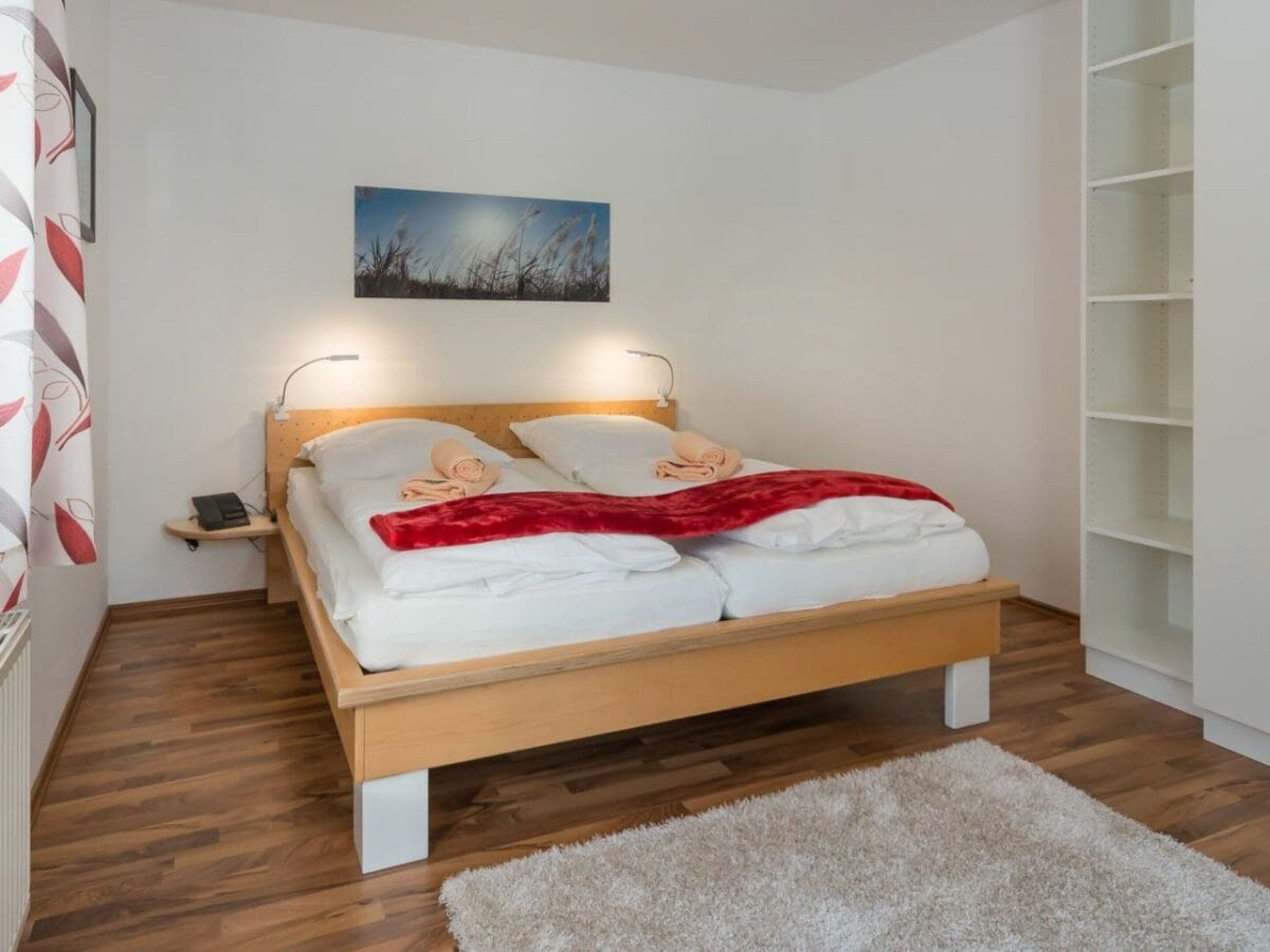 Una cama bien hecha con sábanas blancas y una colcha roja, una mesita de noche con una lámpara, y un cuadro en la pared.