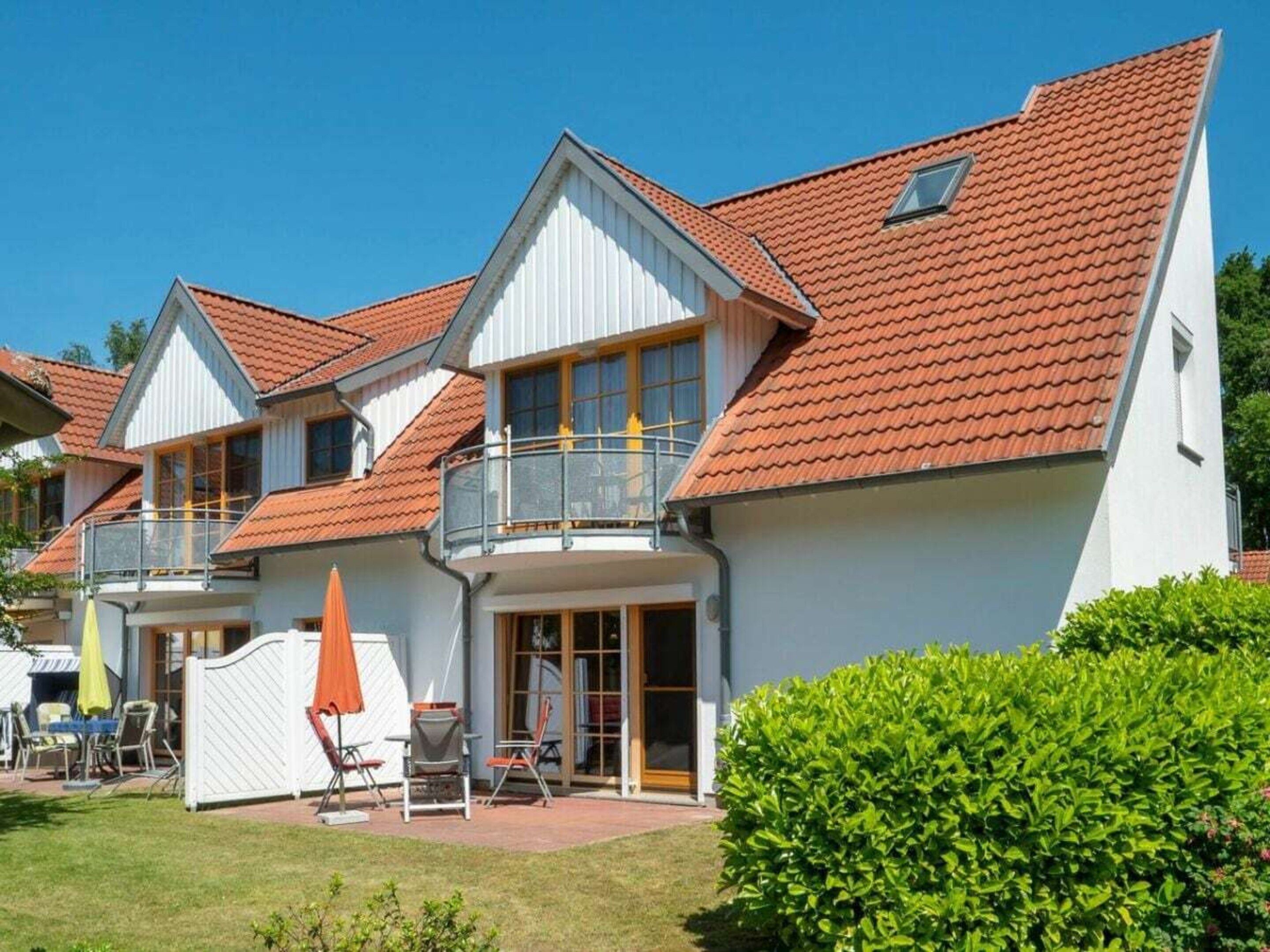 Ein zweigeschossiges Wohngebäude mit rotem Ziegeldach, einem Balkon und einer Terrasse mit Gartenmöbeln.