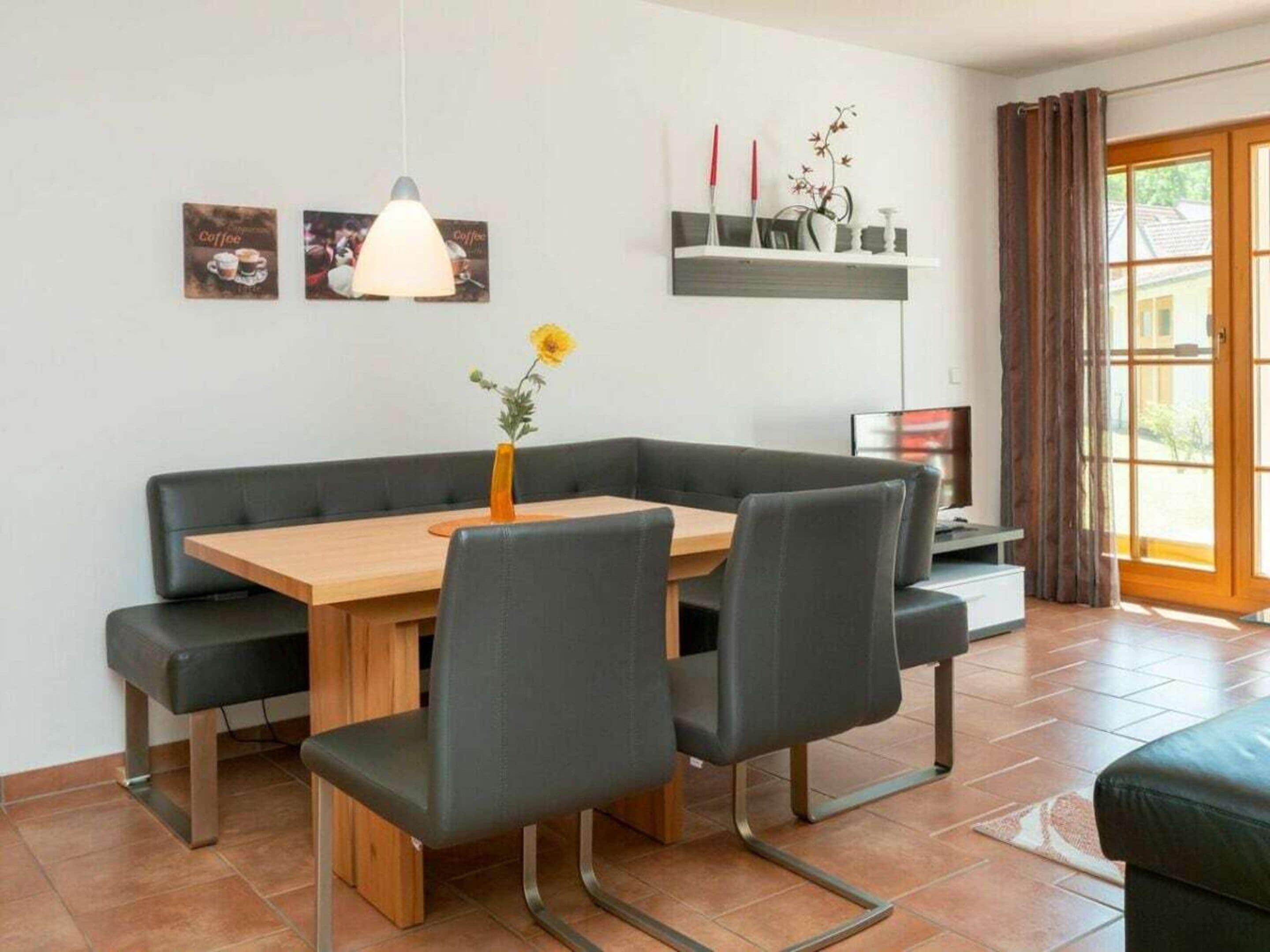 Een moderne eetruimte met een houten tafel, zwarte stoelen met bekleding, een hanglamp en een vaas met een bloem.
