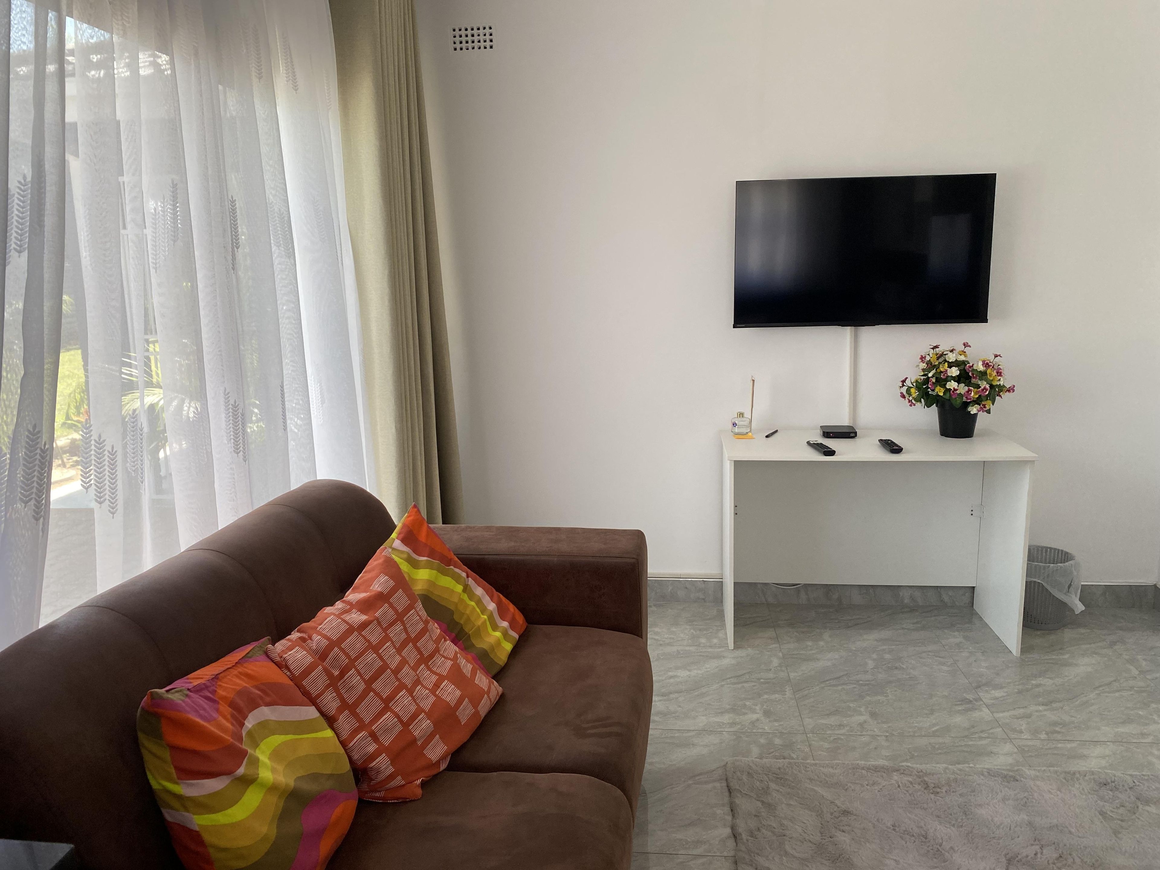 Un salon avec un canapé marron, un bureau blanc, un téléviseur à écran plat et une plante en pot.