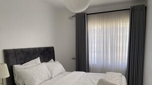 Un lit bien fait, avec du linge de lit blanc et une tête de lit sombre, dans une pièce dotée d’une grande fenêtre et de rideaux.