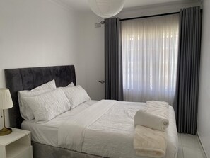 Una cama bien hecha con sábanas blancas y un cabecero oscuro en una habitación con un ventanal y cortinas.