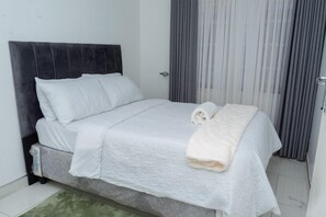 Ein ordentlich bezogenes Bett mit dunklem Kopfteil, weißer Bettwäsche und einem aufgerollten Handtuch an der Seite.