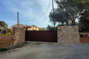 A gated entrance with a stone wall and a metal gate, leading to a property with a house in the background. - Can Costals Olivo in Pals (Masos de Pals)
