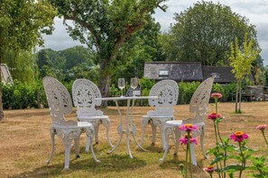 An outdoor seating area with white chairs and a table, surrounded by trees and a building in the background. - The Old Smithy, 2 bedroom apartment - Pass the Keys (Shropshire)