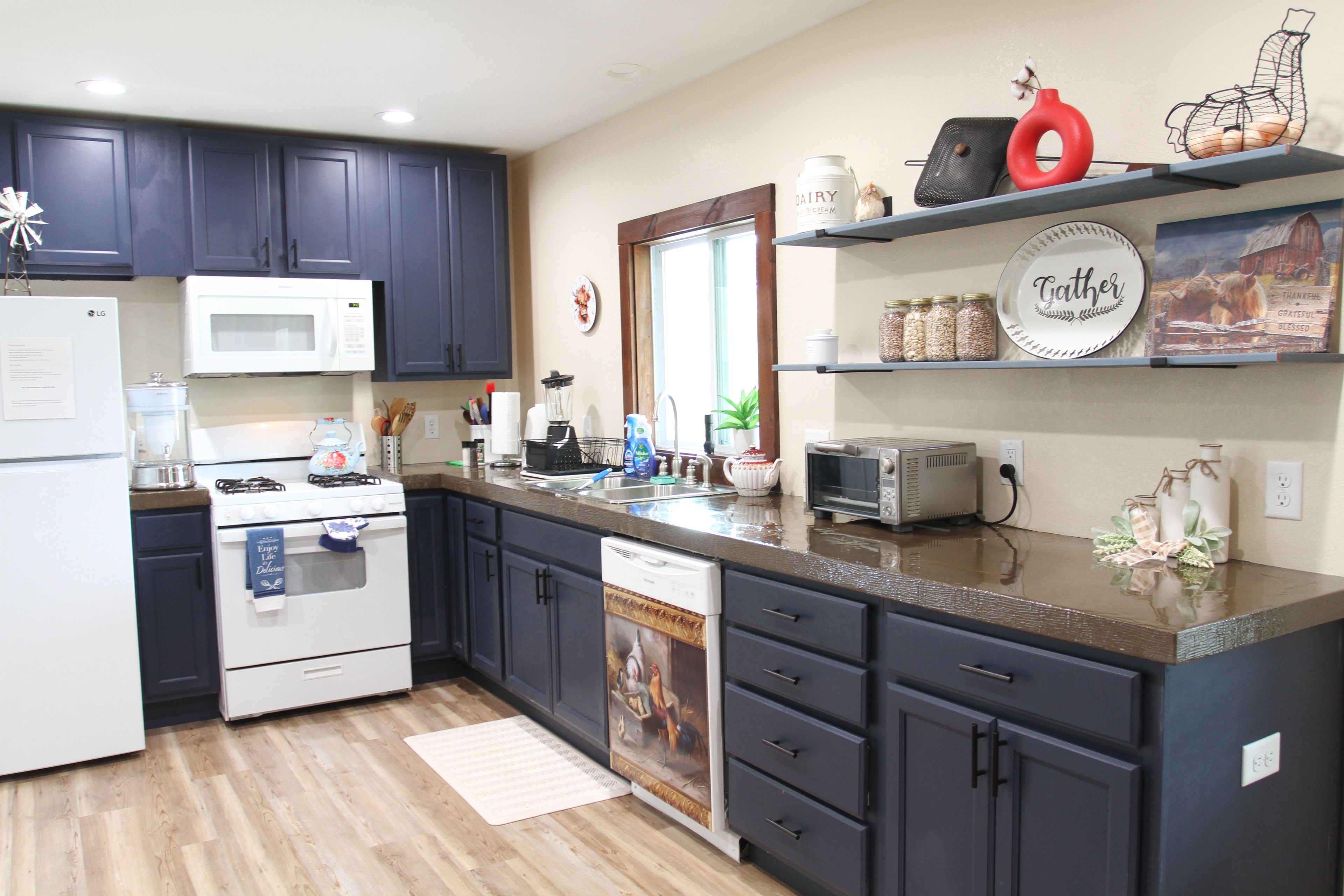 A modern kitchen with dark cabinets, a microwave, stove, and a dishwasher. There are decorative items on the shelves and a window with a view.