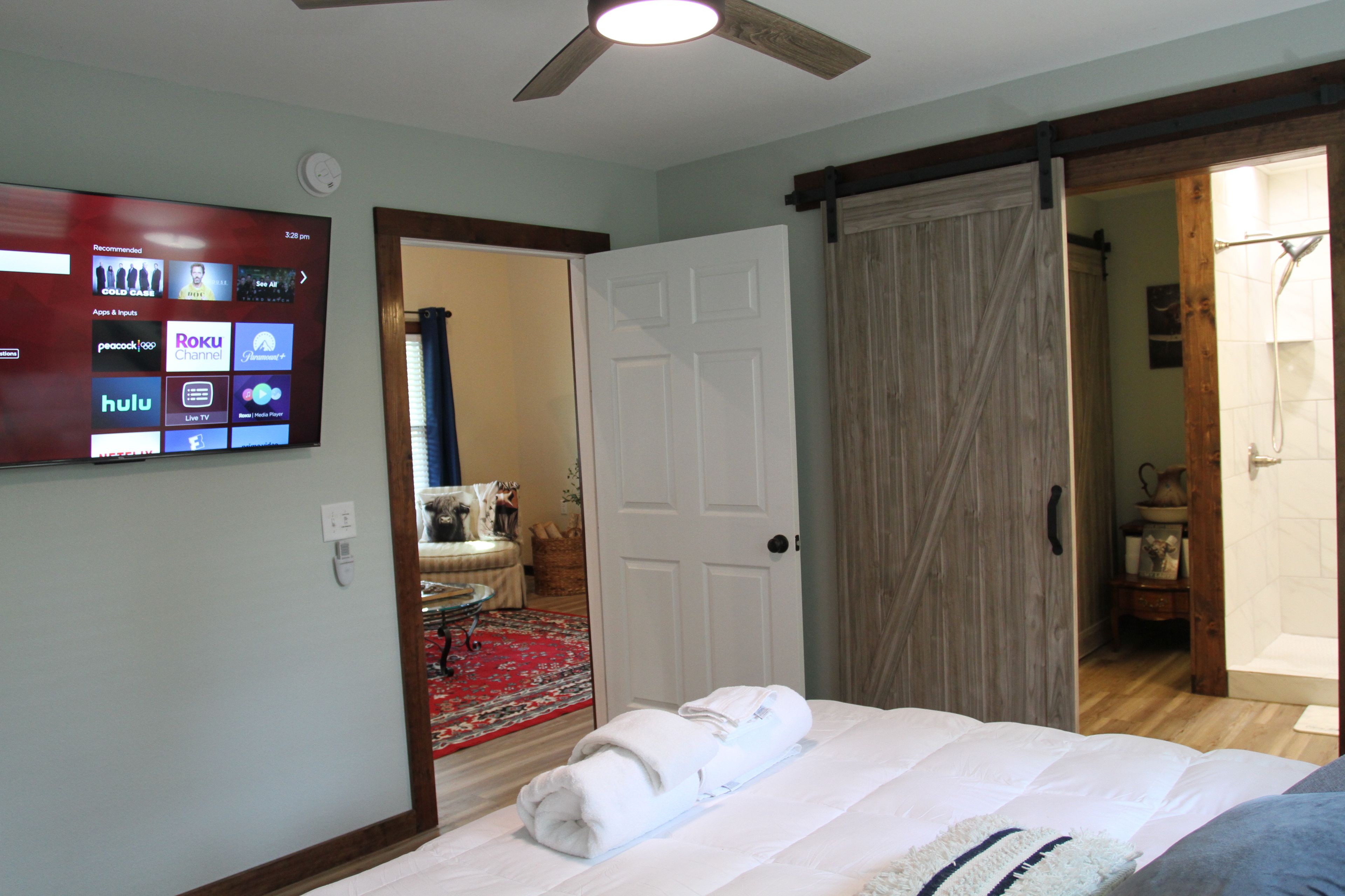 A hotel room with a bed, a television displaying streaming services, a door leading to a bathroom, and a ceiling fan.