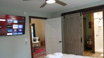 A hotel room with a bed, a television displaying streaming services, a door leading to a bathroom, and a ceiling fan.