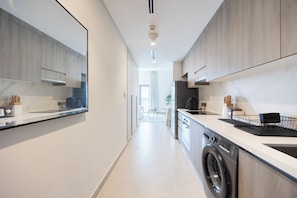 A modern kitchen with a built-in washer and dryer, a large window, and a view into the dining area. - Bloomfields Studio in Albatross 1 - YGC (Abu Dhabi)