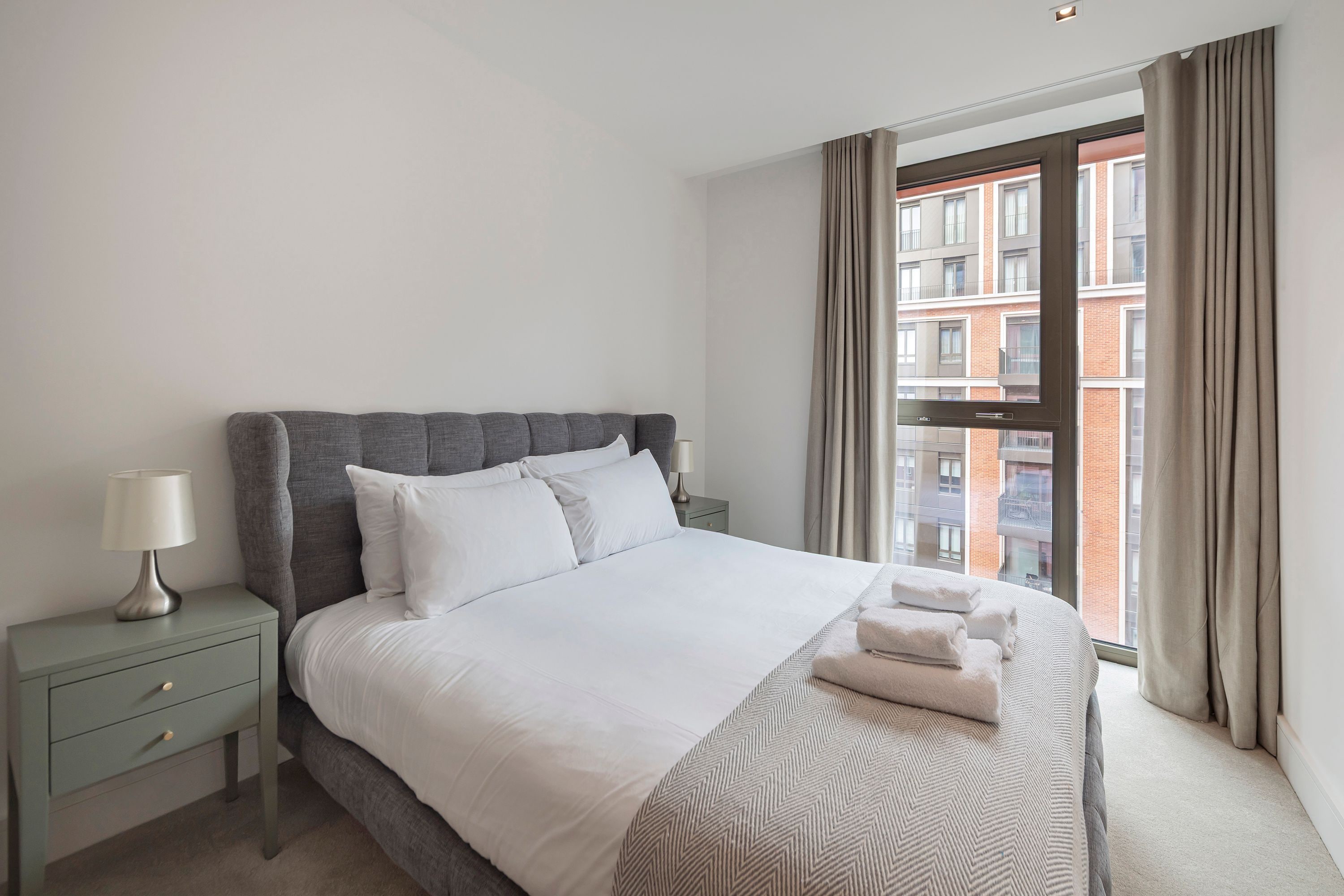 A hotel room with a bed, a nightstand, a lamp, and a view of a building through the window.