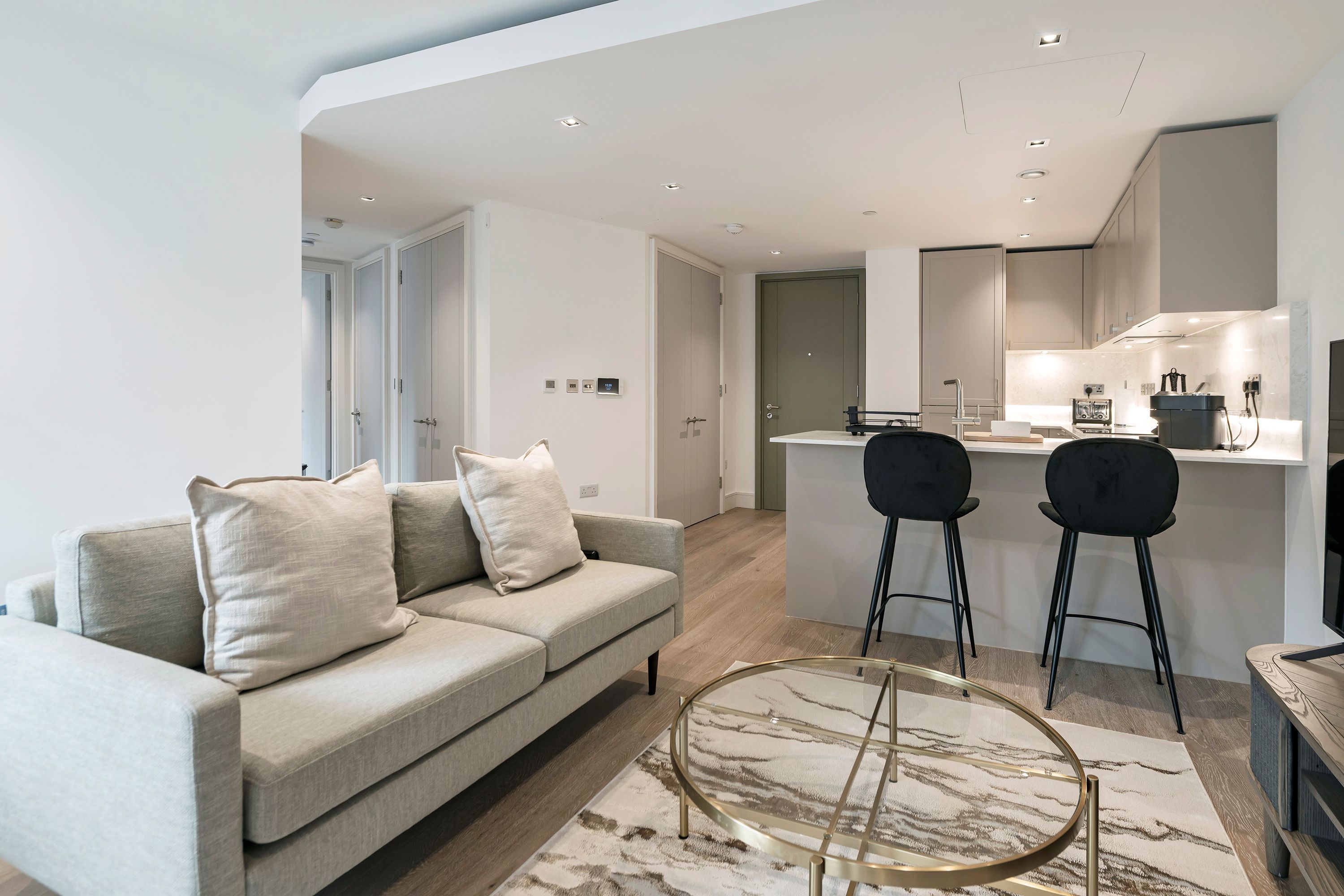 Una sala moderna con un sofá, dos sillones, una mesa de centro de vidrio y una zona de cocina con una mesa de comedor.