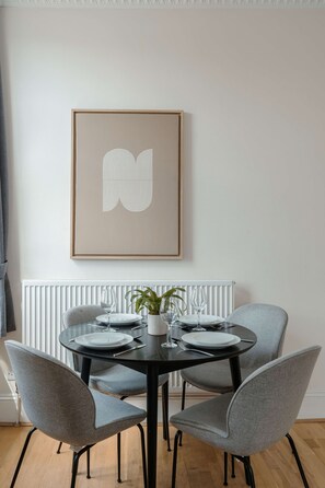 Ein Essbereich mit einem runden Tisch, gedeckt für vier Personen, umgeben von grauen, gepolsterten Stühlen. Darüber hängt ein gerahmtes Kunstwerk mit einem weißen, abstrakten Design.