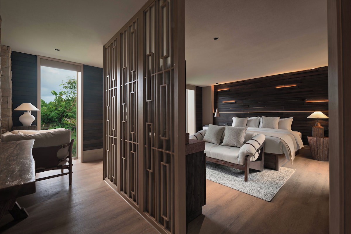 Quarto moderno com uma cama grande, área de estar com uma poltrona e vista para o verde lá fora.