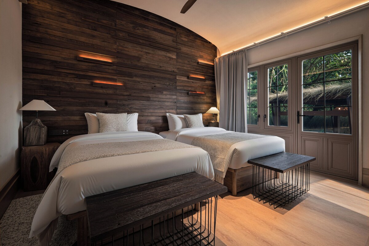 Quarto com duas camas, cabeceira de madeira, luminárias de cabeceira, um banquinho e uma janela grande com vista para árvores.
