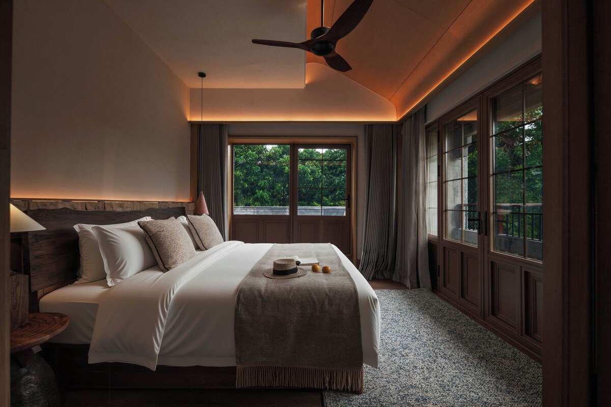 Quarto de hotel com cama, ventoinha no teto, janela grande e vista para o verde lá fora.