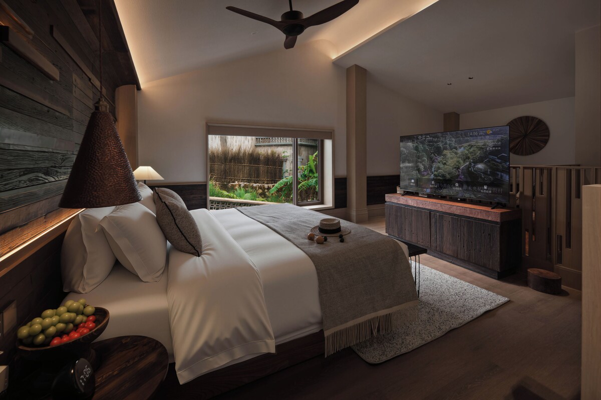 Quarto moderno com uma cama grande, televisão e vista para o verde lá fora.