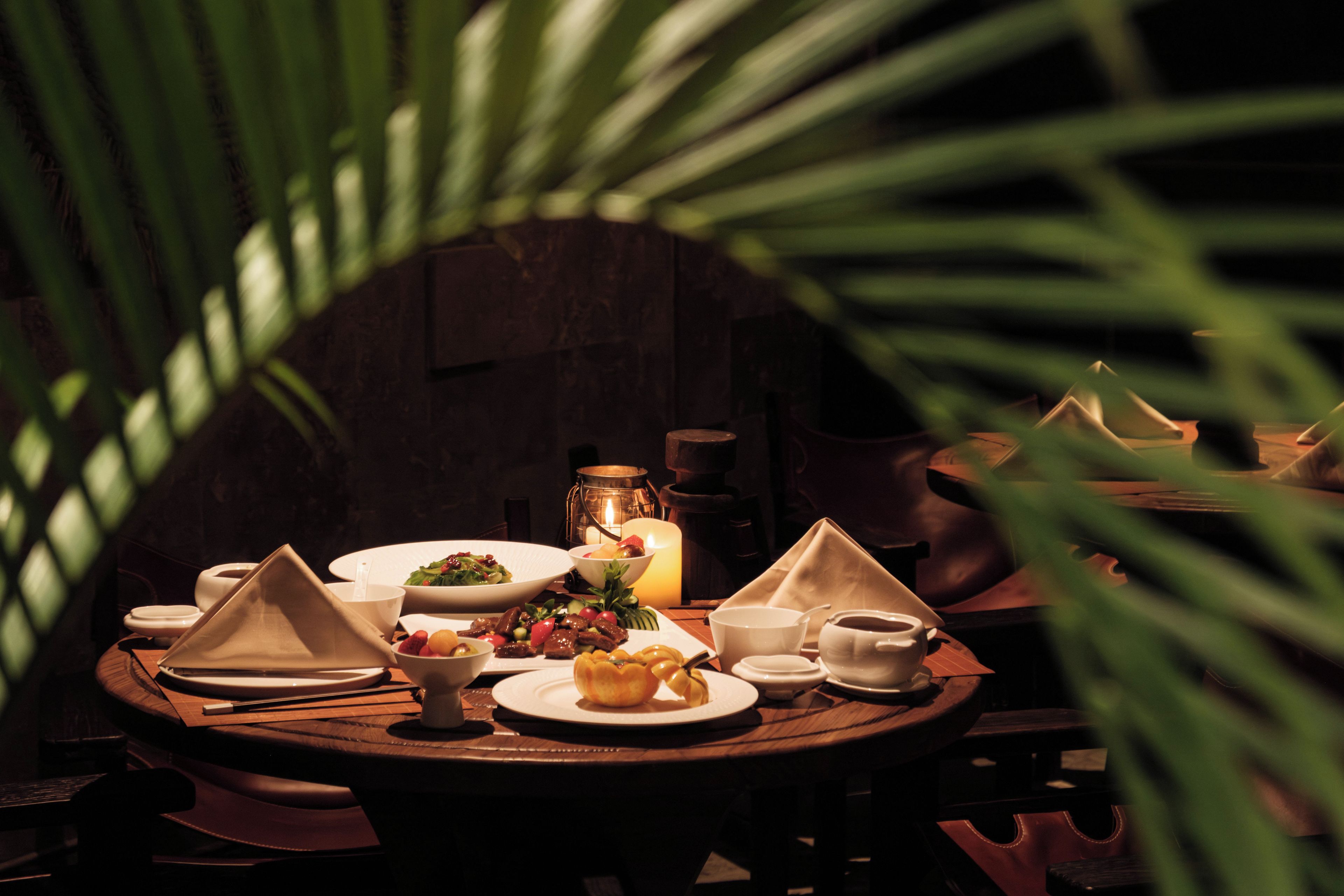 Una mesa de comedor dispuesta con varios platos, una vela y un farol, en un interior oscuro.