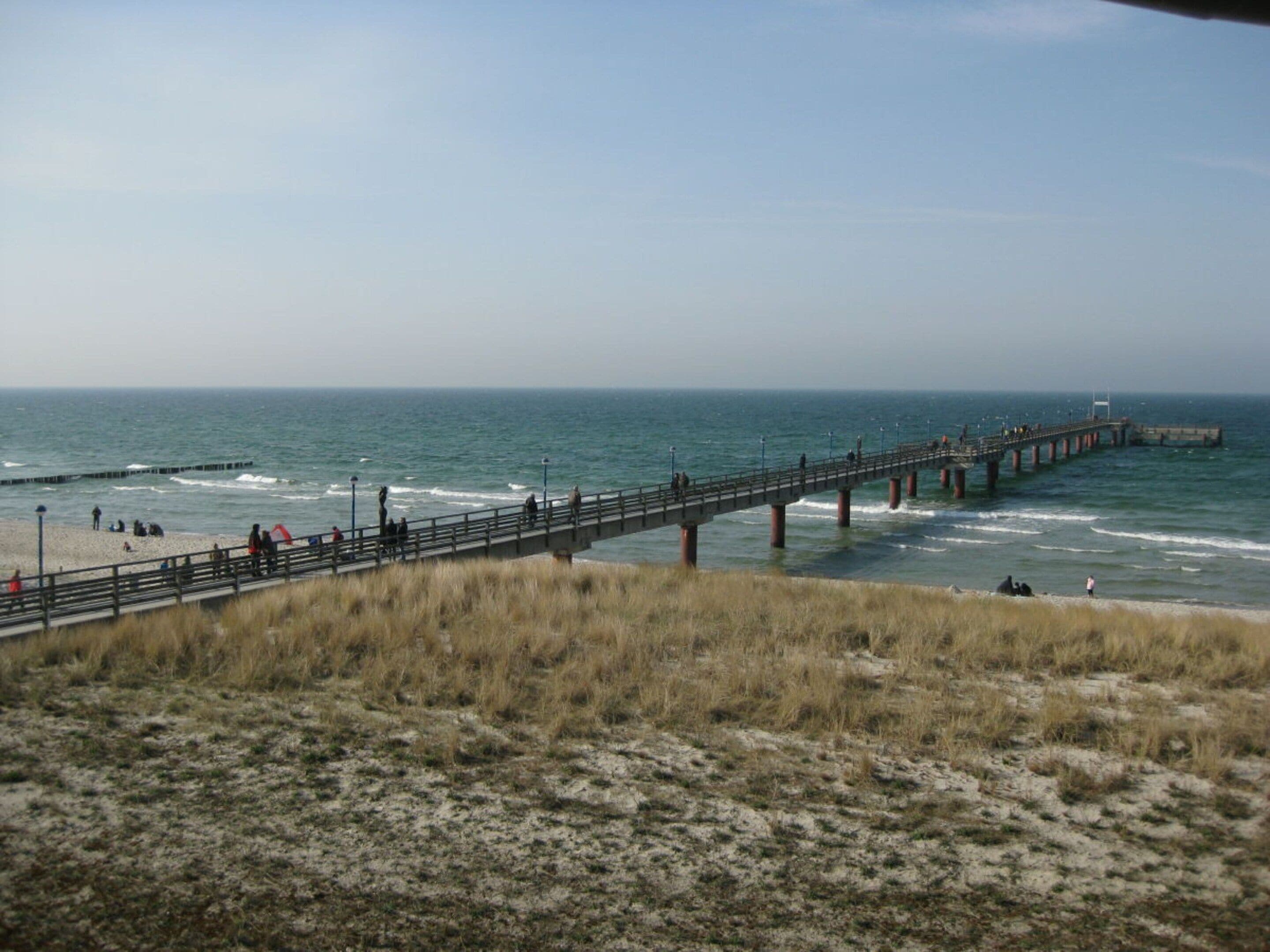 A coastal scene with a pier extending into the sea, sandy beach, and grassy dunes.