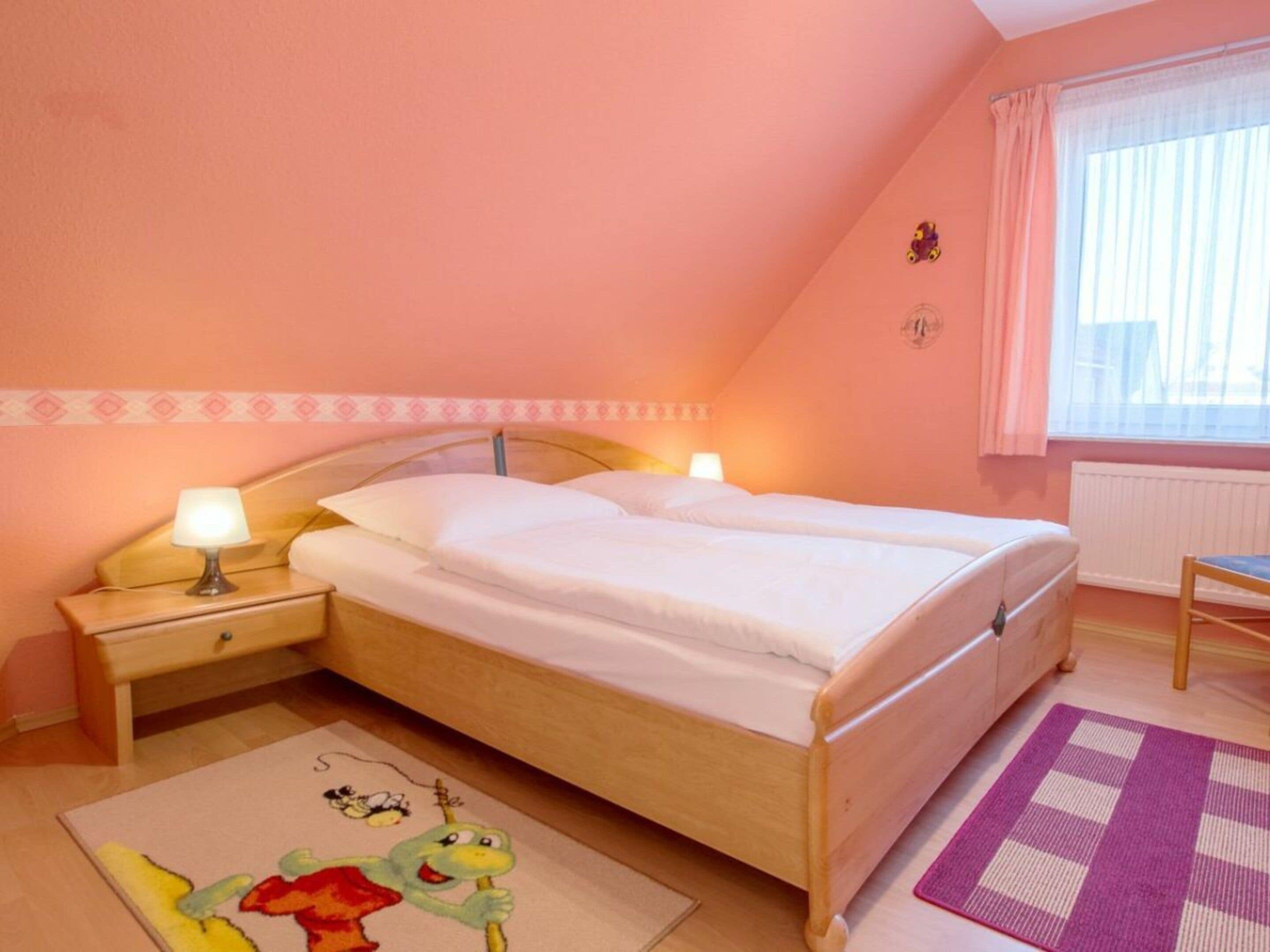 Un letto singolo in una stanza con una testiera in legno, un comodino con una lampada, una finestra con tende e un piccolo tappeto con disegni di personaggi dei cartoni animati.