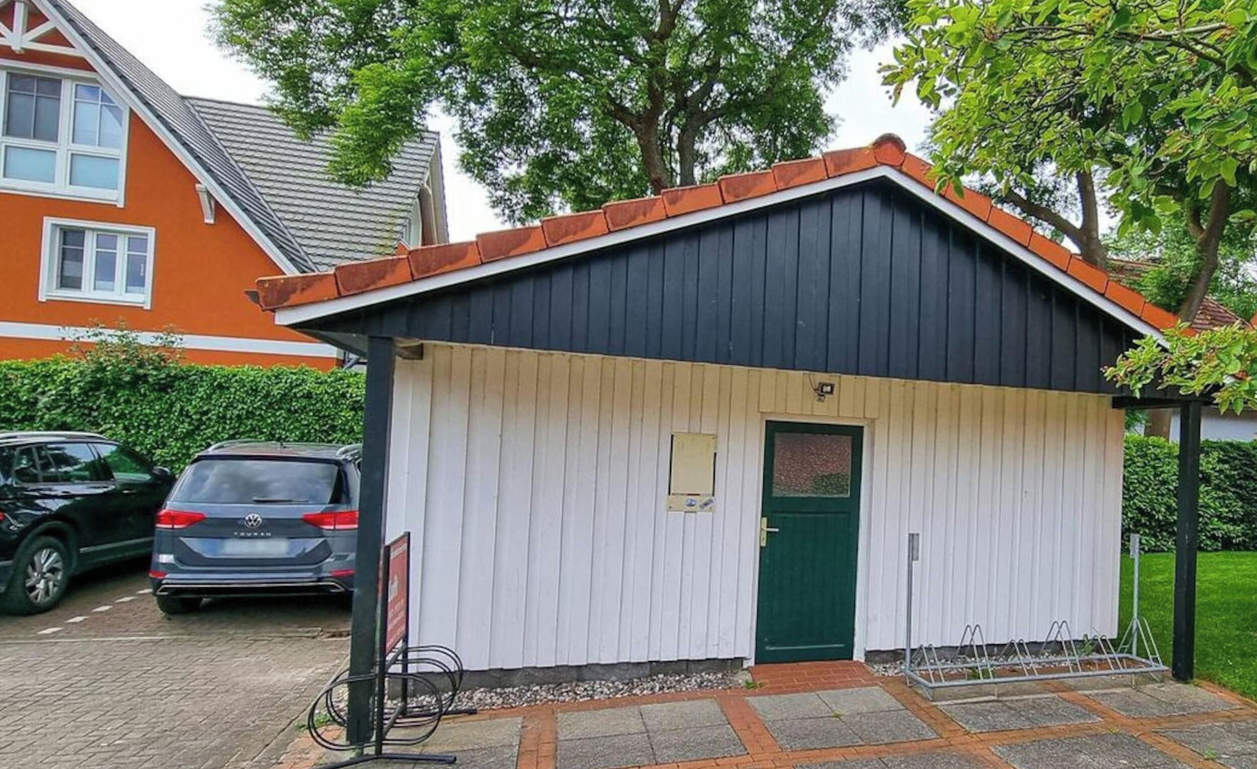 Un piccolo edificio a un solo piano con tetto scuro e pareti bianche, una porta verde e una piccola finestra. Davanti, un'area pavimentata e, a destra, una struttura metallica.