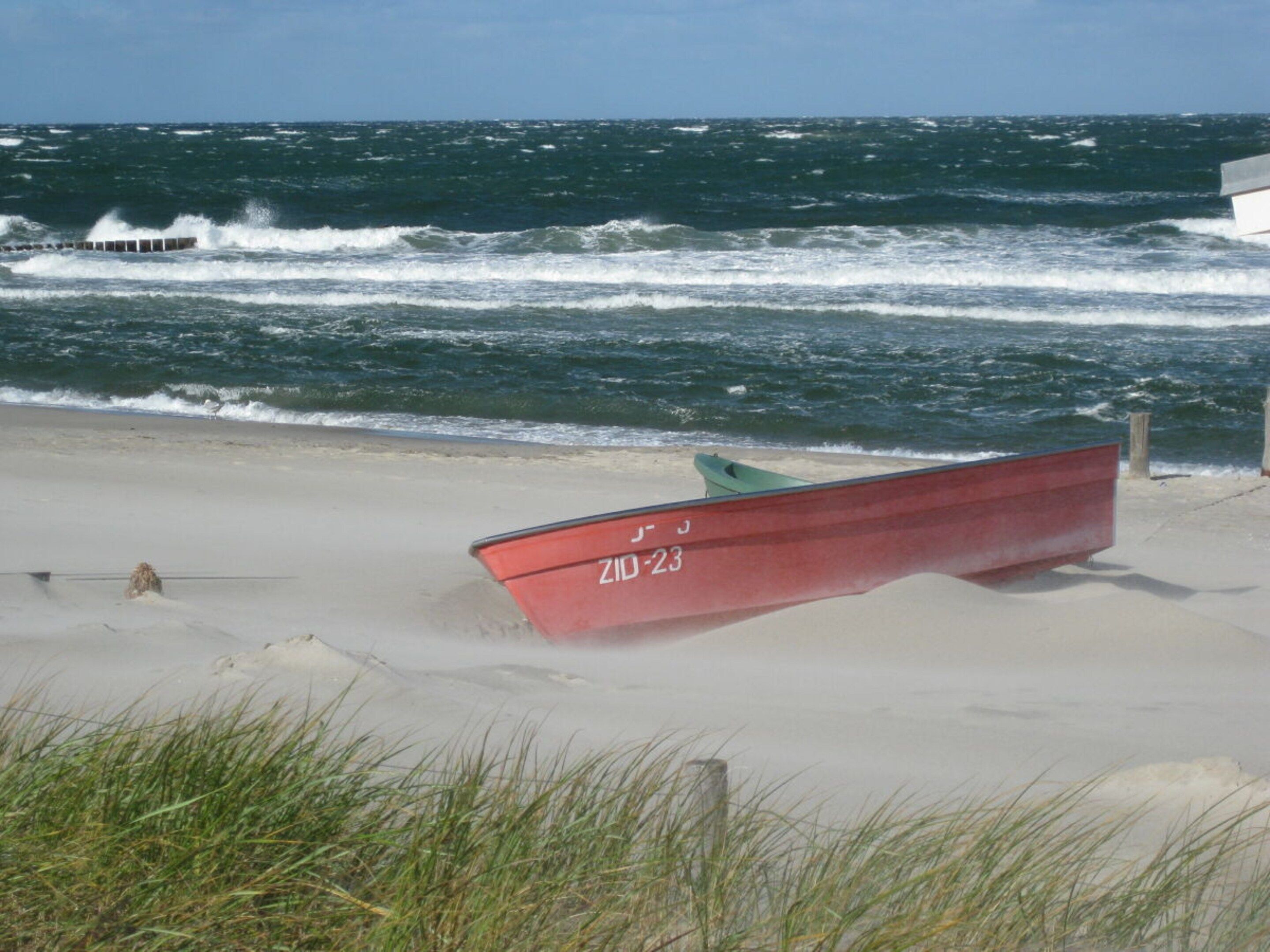 Ein rotes Boot mit der Nummer 'ZID-23' an einem sandigen Strand, im Hintergrund sind Wellen zu sehen.