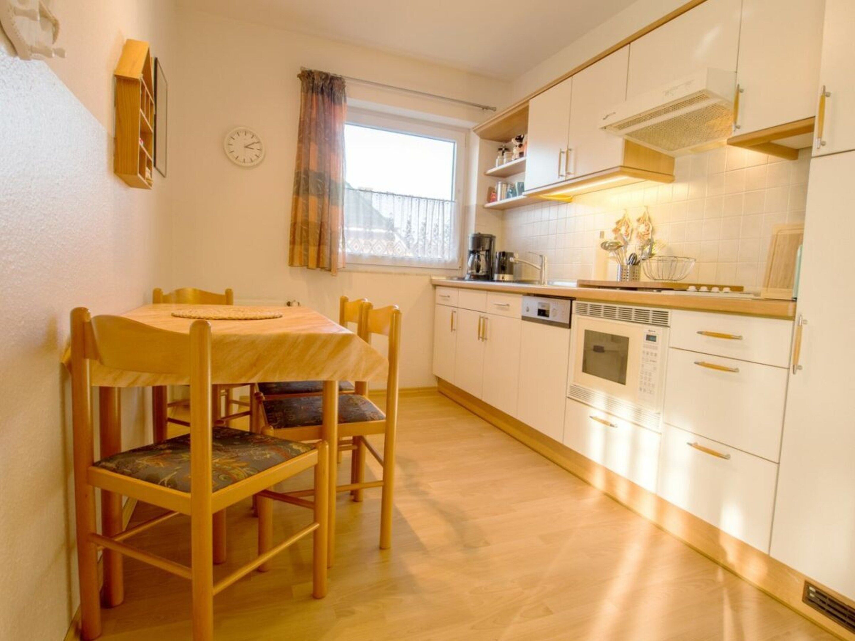 A compact kitchen with white cabinets, a dining table with chairs, a window with curtains, and a clock on the wall.
