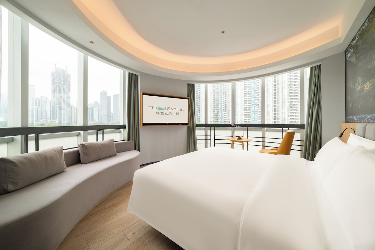 A modern hotel room with a large bed, a seating area with cushions, a small table, and a city view through large windows.