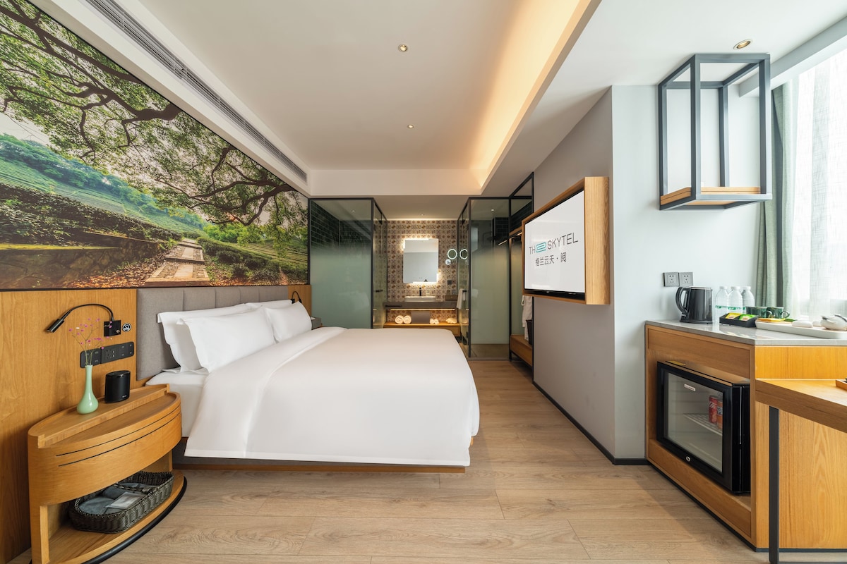 A modern hotel room with a large bed, a desk with a coffee maker, a TV, and a view of a green landscape through the window.