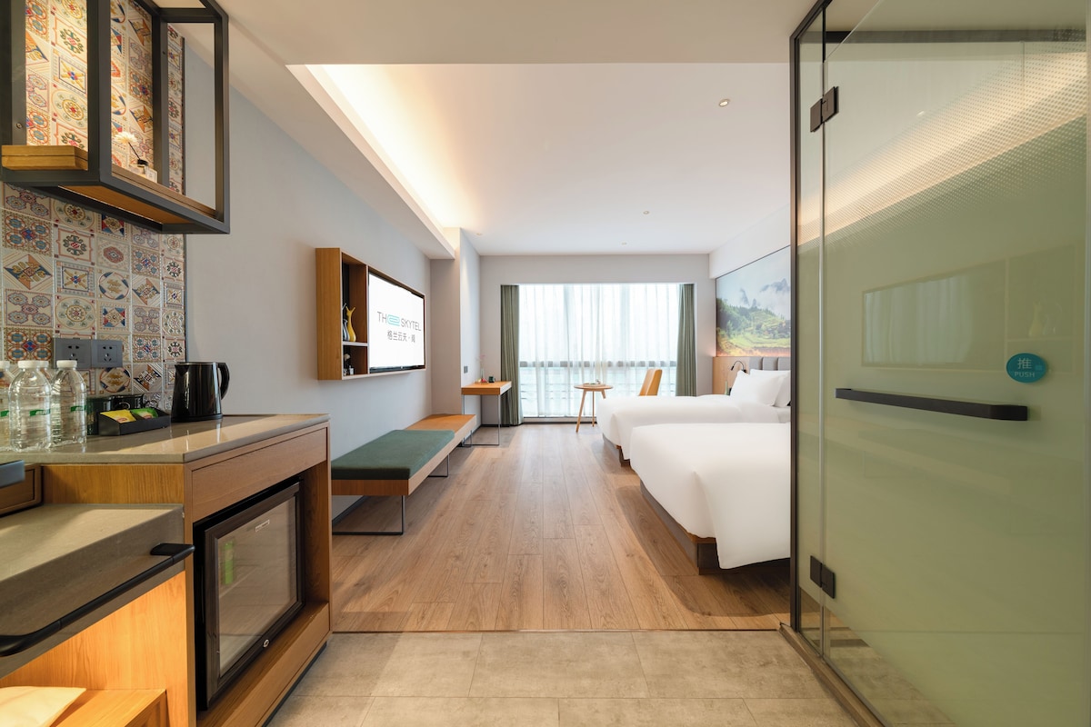 A hotel room with a bed, a desk, a seating area, a minibar, and a view of a mountainous landscape.