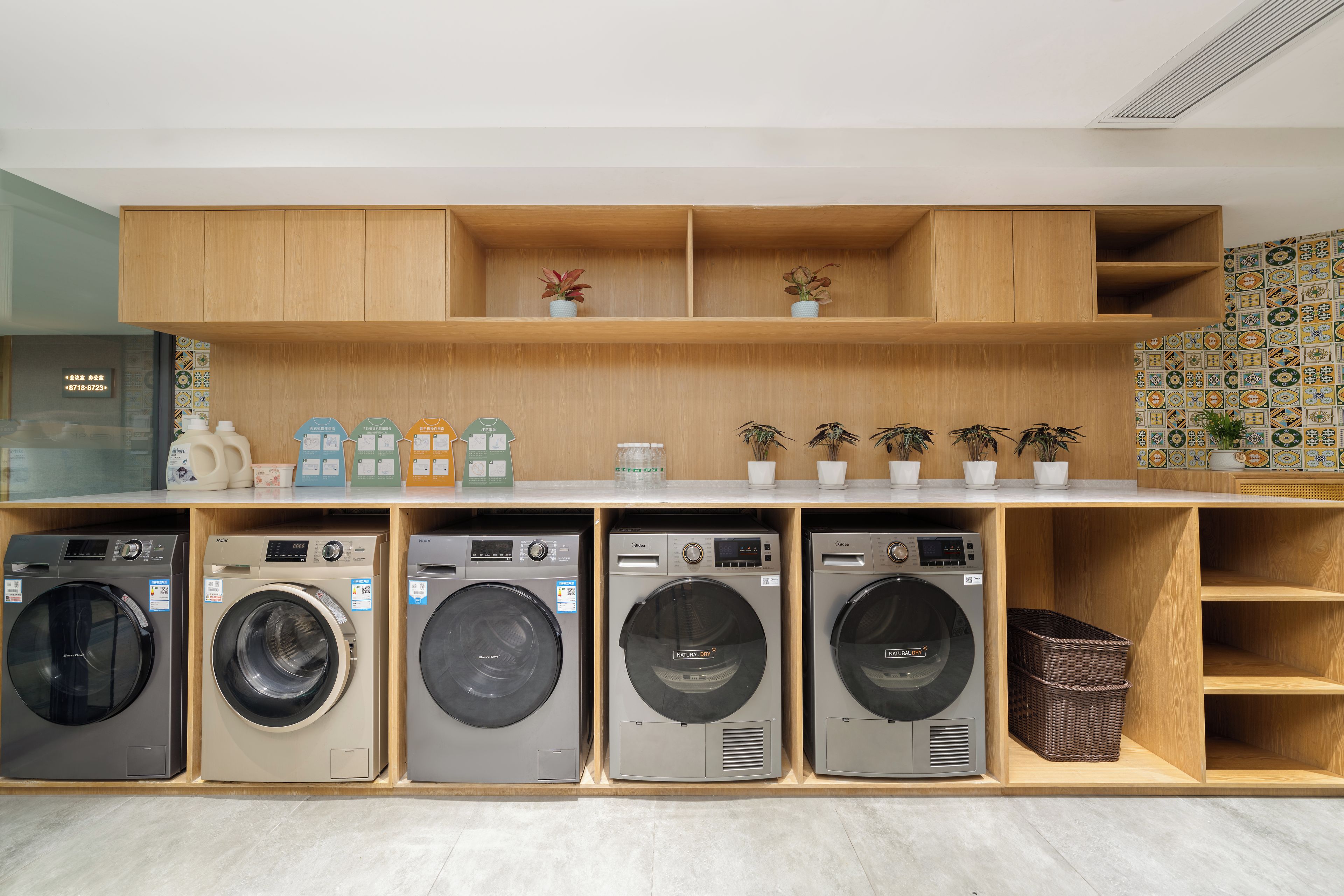 A laundry facility with five front-loading washing machines, wooden shelving units, and decorative potted plants.